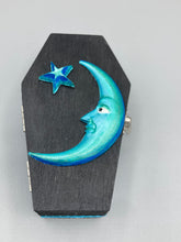 Load image into Gallery viewer, This sleek black box has an eye-catching turquoise crescent at its center, accompanied by a bright blue ribbon and a dainty white moon. Inside, the interior is decorated with a vivid royal blue and detailed with a small crescent moon and black-and-silver cross. Coffin Dimensions: Length: 4 1/2 Inches; Width: 2 1/2 inches; Height: 1 5/8 Inches