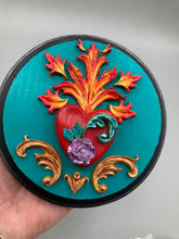 Load image into Gallery viewer, This Sacred Heart Wall Art Decor is the perfect way to enhance your home with an artistic flair. This beautiful artwork features a round bright turquoise wooden frame with a hand-crafted bright red heart. The heart is detailed with light purple and vibrant green leaves, with a vibrant fire embellishment over the center. Gold decorative embellishments at the sides of the heart add a luxurious touch. Diameter 4"