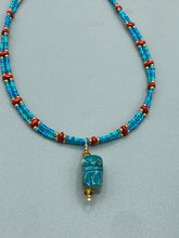Load image into Gallery viewer, This adjustable choker style necklace features an eye-catching teal wood scarab beetle pendant and glass beaded strands. This dramatic necklace is kept together by double-hole flat matte lava-red beads, providing a collar shape. The necklace artfully wraps around the neck, conveying an elegant and bold style.  Measurements:    Necklace is 14 1/2” in length, can be adjusted up to 16"