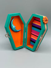 Load image into Gallery viewer, This coffin-shaped box features a bright turquoise exterior, with a bold yellow, orange and red sun adorning the center. Complemented with a shimmery rose-gold ribbon and flower, the inside is painted in a vibrant orange hue with turquoise crescent moon detailing and traditional Mexican-themed fabric. Coffin Dimensions: Length: 4 1/2 Inches; Width: 2 1/2 inches; Height: 1 5/8 Inches