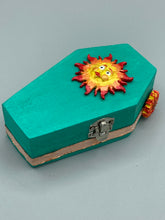 Load image into Gallery viewer, This coffin-shaped box features a bright turquoise exterior, with a bold yellow, orange and red sun adorning the center. Complemented with a shimmery rose-gold ribbon and flower, the inside is painted in a vibrant orange hue with turquoise crescent moon detailing and traditional Mexican-themed fabric. Coffin Dimensions: Length: 4 1/2 Inches; Width: 2 1/2 inches; Height: 1 5/8 Inches