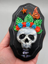 Load image into Gallery viewer, Showcasing a vibrant Frida Kahlo inspired calavera with a dark wood frame, this Day of the Dead wall decor piece is perfect for adding a traditional Mexican folk art touch to any home. Featuring an intricately detailed clay calavera in the style of Frida Kahlo, this Day of the Dead wall art decor is sure to be the centerpiece of any home décor. The calavera is wearing Frida's iconic crown of flowers and a monarch butterfly, ensuring it will bring both a unique and inspiring look to any wall.