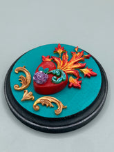 Load image into Gallery viewer, This Sacred Heart Wall Art Decor is the perfect way to enhance your home with an artistic flair. This beautiful artwork features a round bright turquoise wooden frame with a hand-crafted bright red heart. The heart is detailed with light purple and vibrant green leaves, with a vibrant fire embellishment over the center. Gold decorative embellishments at the sides of the heart add a luxurious touch. Diameter 4"