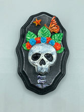 Load image into Gallery viewer, Showcasing a vibrant Frida Kahlo inspired calavera with a dark wood frame, this Day of the Dead wall decor piece is perfect for adding a traditional Mexican folk art touch to any home. Featuring an intricately detailed clay calavera in the style of Frida Kahlo, this Day of the Dead wall art decor is sure to be the centerpiece of any home décor. The calavera is wearing Frida's iconic crown of flowers and a monarch butterfly, ensuring it will bring both a unique and inspiring look to any wall.