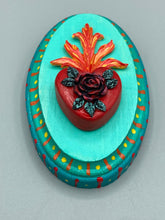 Load image into Gallery viewer, This Sacred Heart Wall Art Decor is the perfect way to enhance your home with an artistic flair. This beautiful artwork features a bright turquoise wooden frame with a hand-crafted bright red heart. The heart is detailed with dark red rose and vibrant green leaves, with a vibrant fire embellishment over the center. Gold decorative embellishments at the sides of the heart add a luxurious touch. H 5 3/8" x W 3 3/8” x D 1 1/2"