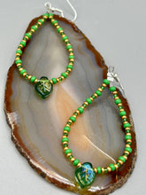 Load image into Gallery viewer, This pair of earrings features a dark green, pressed glass leaf charm in an iridescent finish that gives off subtle hues of gold, purple, blue, and pink. Complemented with green and gold glass beaded hoop earrings, this set is sure to sparkle.
