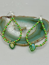 Load image into Gallery viewer, This pair of earrings features a dark green, pressed glass leaf charm in an iridescent finish that gives off subtle hues of gold, purple, blue, and pink. Complemented with green and gold glass beaded hoop earrings, this set is sure to sparkle.
