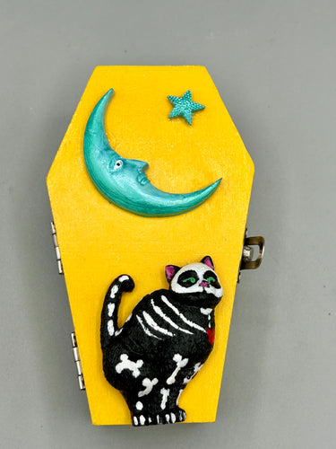 Coffin keepsake box represents the eternal love for a special kitty cat in your life. Cat silhouette sitting admiring the moon and stars. Day Of The Dead art was created for the cat lovers who adore their sweet kitty. Coffin-shaped box features a bright and vibrant canary yellow exterior, with an adoring blue crescent moon and star. Complemented with a shimmery blue ribbon and flower, the inside is painted in a bright red hue with black floral cross and detailing a traditional Mexican-themed fabric.