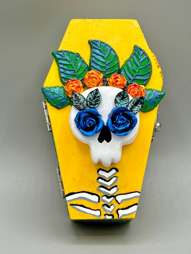 This coffin-shaped box features a bright canary yellow exterior, showcasing a vibrant Frida Kahlo inspired calavera. This beautifully handcrafted calavera is adorned with Frida's iconic floral crown. Complemented with a shimmery pastel-blue ribbon, the inside is painted in a bright blue hue accompanied by a black and silver floral cross and a Day Of The Dead kitten. The interior is also finished with a traditional Mexican-themed fabric.