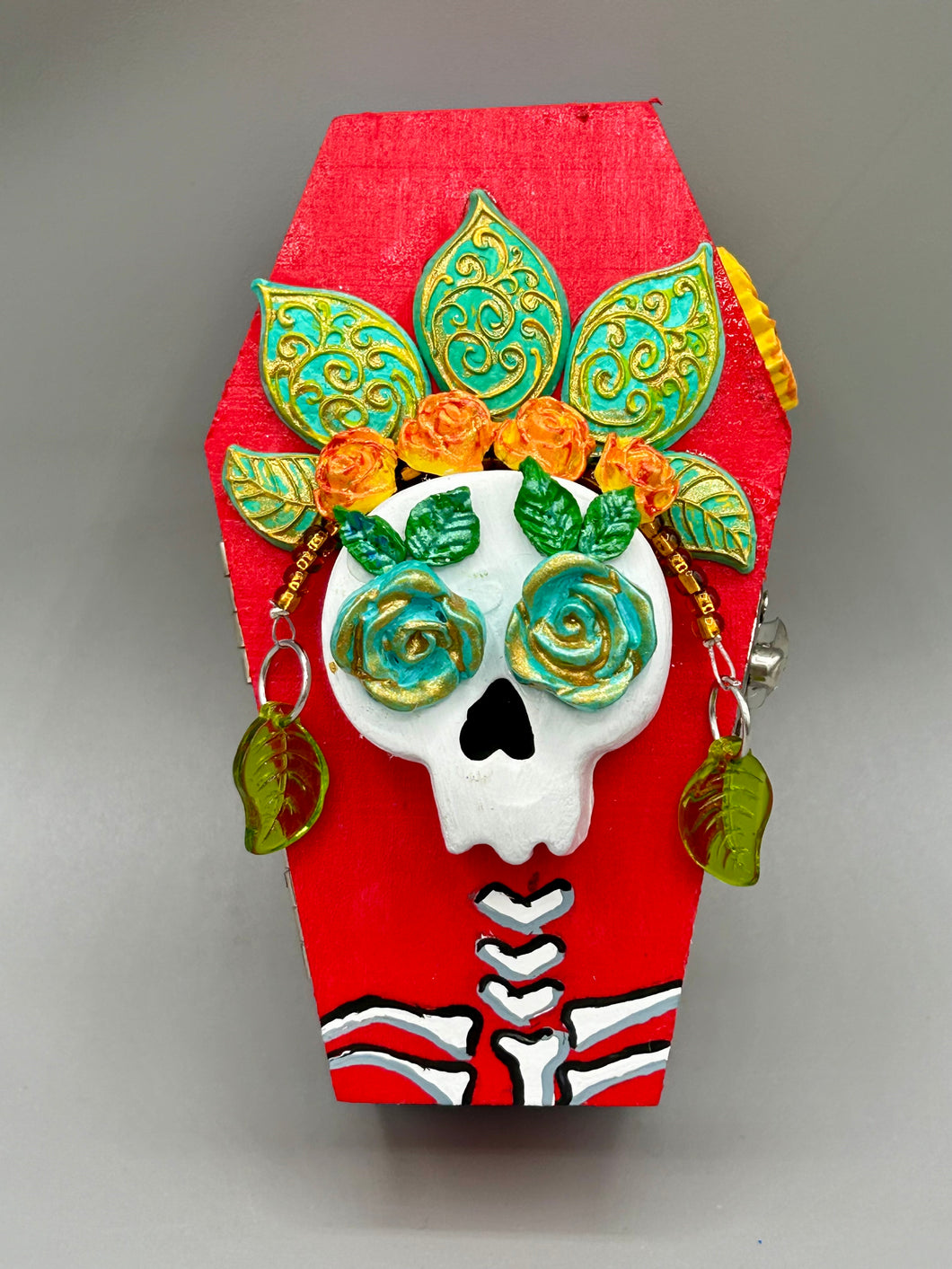 This coffin-shaped box features a bright and colorful exterior, showcasing a vibrant Frida Kahlo inspired calavera.  This beautifully handcrafted calavera is adorned with  Frida's iconic floral crown. To complete the look, the inside is also painted in colorful hues with a black and silver floral cross. The interior is also finished with a traditional Mexican-themed fabric.  L: 4 1/2“; W: 2 ½”; H: 1 5/8”