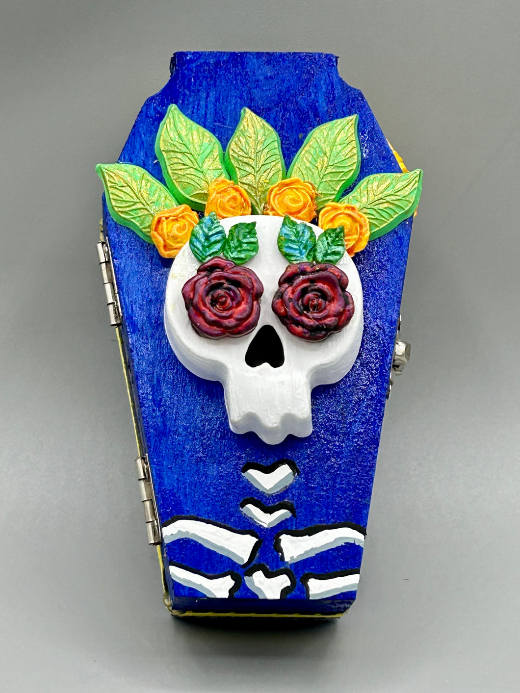This coffin-shaped box features a bright and colorful exterior, showcasing a vibrant Frida Kahlo inspired calavera.  This beautifully handcrafted calavera is adorned with  Frida's iconic floral crown. To complete the look, the inside is also painted in colorful hues with a black and silver floral cross. The interior is also finished with a traditional Mexican-themed fabric.  L: 4 1/2“; W: 2 ½”; H: 1 5/8”