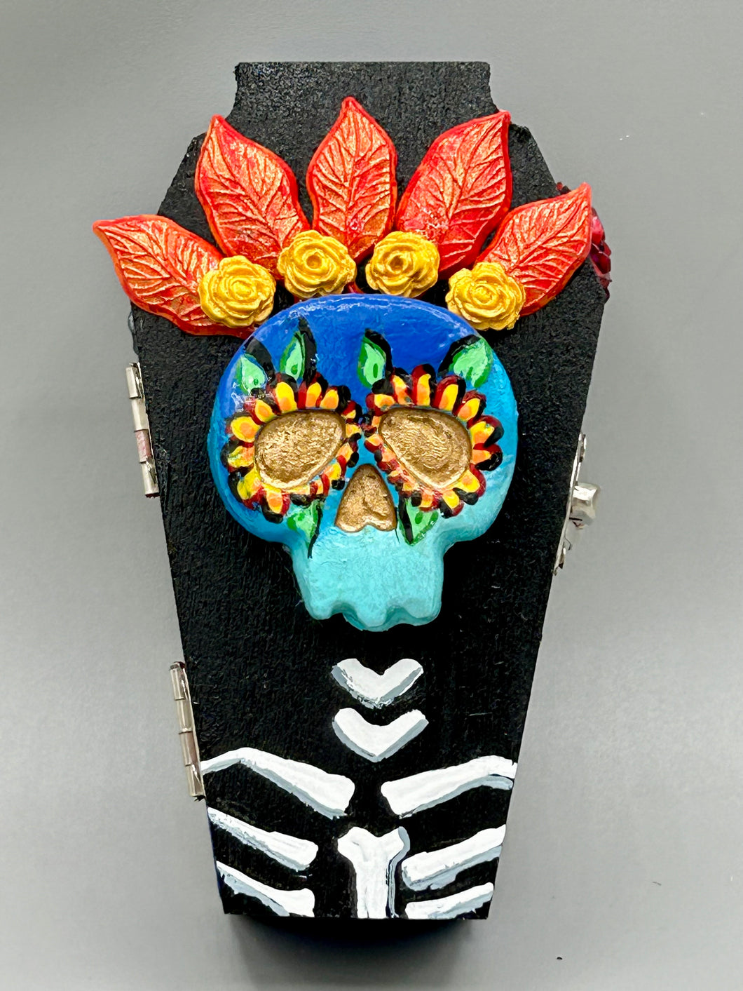 This coffin-shaped box features a bright and colorful exterior, showcasing a vibrant Frida Kahlo inspired calavera.  This beautifully handcrafted calavera is adorned with  Frida's iconic floral crown. To complete the look, the inside is also painted in colorful hues with a black and silver floral cross. The interior is also finished with a traditional Mexican-themed fabric.  L: 4 1/2“; W: 2 ½”; H: 1 5/8”