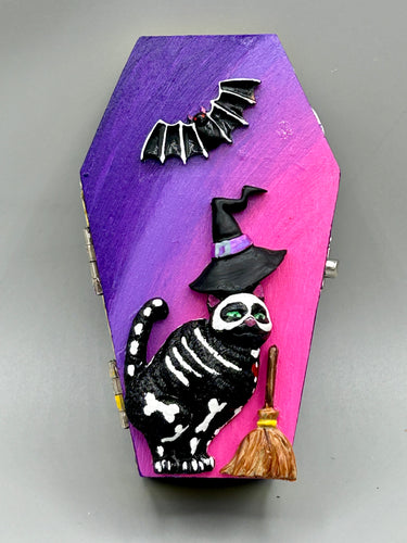 Coffin shaped box features a Day of the Dead kitty cat wearing a cute witchy hat while standing next to her witchy broom and a flying bat friend.  The inside of the coffin is adorned with a witchy crescent moon, Jack-O-Lantern and a cute bat. This captivating coffin box makes an eye-catching, bewitching and whimsical Halloween-themed art piece that adds a one-of-a-kind Halloween touch to any home or office.  L: 4 7/8