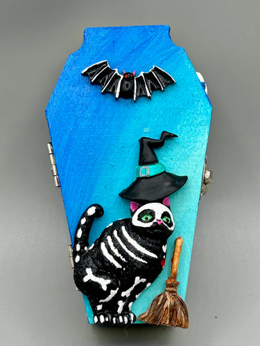 Coffin shaped box features a Day of the Dead kitty cat wearing a cute witchy hat while standing next to her witchy broom and her flying bat friend. The inside of the coffin is adorned with a witchy crescent moon, Jack-O-Lantern and a cute Day of the Dead tiny kitten. This captivating coffin box makes an eye-catching, bewitching and whimsical Halloween-themed art piece that adds a one-of-a-kind Halloween touch to any home or office.