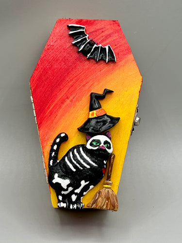 Coffin shaped box features a Day of the Dead kitty cat wearing a cute witchy hat while standing next to her witchy broom and her flying bat friend. The inside of the coffin is adorned with a witchy crescent moon, Jack-O-Lantern and a cute bat. This captivating coffin box makes an eye-catching, bewitching and whimsical Halloween-themed art piece that adds a one-of-a-kind Halloween touch to any home or office.