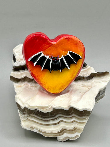 This hand-crafted art magnet is featuring a black bat with an ombre background of yellow, orange and bright red. This art clay magnet is the ideal home decor for Halloween enthusiasts. Perfect for adorning a witchy altar, goth, or spooky Halloween home decor.  Halloween magnet art to add to your fridge, locker, cubicle or toolbox. This hand-crafted heart-shaped magnet features a black bat figurine with ombre colors of yellow, orange and bright red.