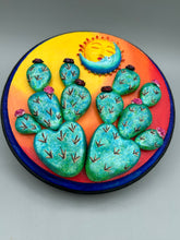Load image into Gallery viewer, Southwest inspired wall art comes in a circle-shaped wood frame with a sun and crescent moon motifs. Its warm sky hues, including yellow, orange and dark red, are further complemented by hand sculpted cacti plants with cool green-blue tones with light and dark pink flowers. Diameter 6 3/4&quot;
