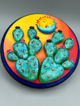 Load image into Gallery viewer, Southwest inspired wall art comes in a circle-shaped wood frame with a sun and crescent moon motifs. Its warm sky hues, including yellow, orange and dark red, are further complemented by hand sculpted cacti plants with cool green-blue tones with light and dark pink flowers. Diameter 6 3/4&quot;

