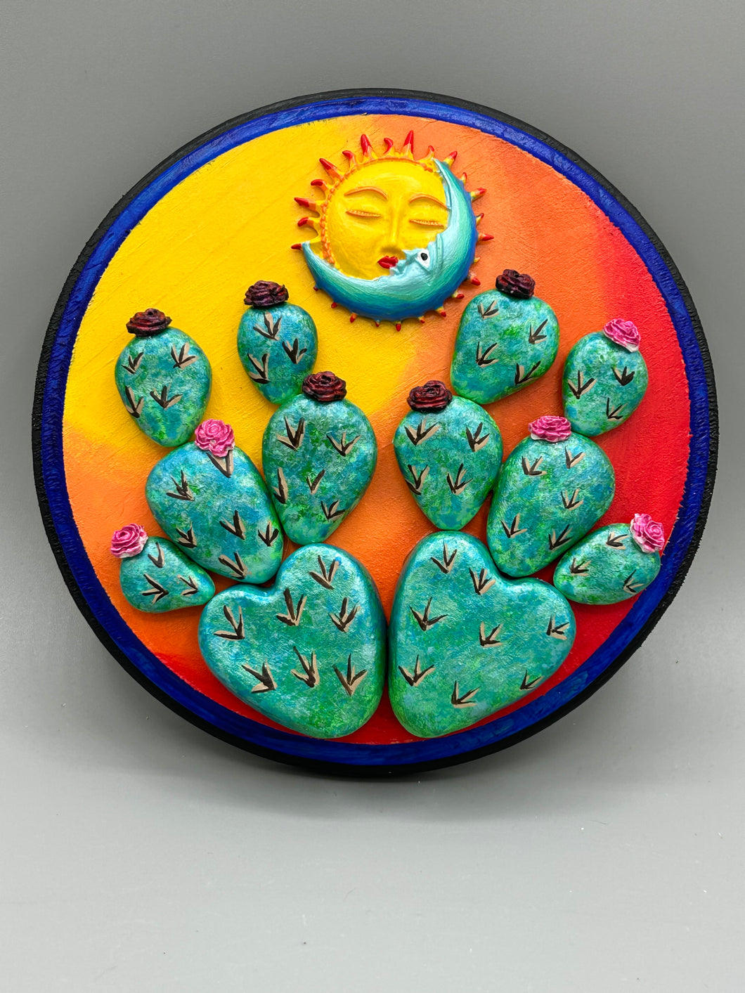 Southwest inspired wall art comes in a circle-shaped wood frame with a sun and crescent moon motifs. Its warm sky hues, including yellow, orange and dark red, are further complemented by hand sculpted cacti plants with cool green-blue tones with light and dark pink flowers. Diameter 6 3/4