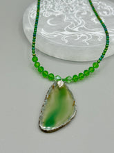 Load image into Gallery viewer, This Light Green Agate Geode Sliced Stone Chip Necklace is a statement piece for your jewelry collection. It features a bold green agate geode pendant surrounded by a glass bead necklace in varying shades of green. The silver leaf bail completes this chic, Boho-inspired necklace.  Measurements:   Necklace is 18” in length - pendant is 2 1/4” Height
