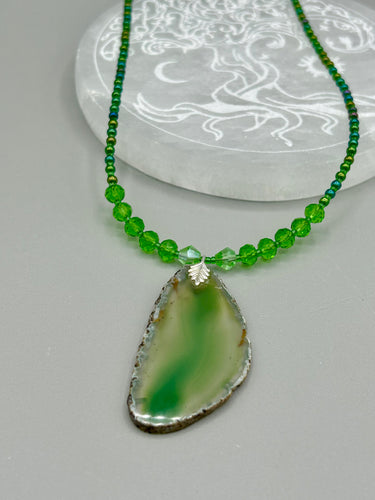 This Light Green Agate Geode Sliced Stone Chip Necklace is a statement piece for your jewelry collection. It features a bold green agate geode pendant surrounded by a glass bead necklace in varying shades of green. The silver leaf bail completes this chic, Boho-inspired necklace.  Measurements:   Necklace is 18” in length - pendant is 2 1/4” Height