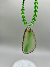 Load image into Gallery viewer, This Light Green Agate Geode Sliced Stone Chip Necklace is a statement piece for your jewelry collection. It features a bold green agate geode pendant surrounded by a glass bead necklace in varying shades of green. The silver leaf bail completes this chic, Boho-inspired necklace.  Measurements:   Necklace is 18” in length - pendant is 2 1/4” Height
