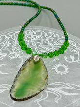 Load image into Gallery viewer, This Light Green Agate Geode Sliced Stone Chip Necklace is a statement piece for your jewelry collection. It features a bold green agate geode pendant surrounded by a glass bead necklace in varying shades of green. The silver leaf bail completes this chic, Boho-inspired necklace.  Measurements:   Necklace is 18” in length - pendant is 2 1/4” Height

