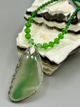 Load image into Gallery viewer, This Light Green Agate Geode Sliced Stone Chip Necklace is a statement piece for your jewelry collection. It features a bold green agate geode pendant surrounded by a glass bead necklace in varying shades of green. The silver leaf bail completes this chic, Boho-inspired necklace.  Measurements:   Necklace is 18” in length - pendant is 2 1/4” Height
