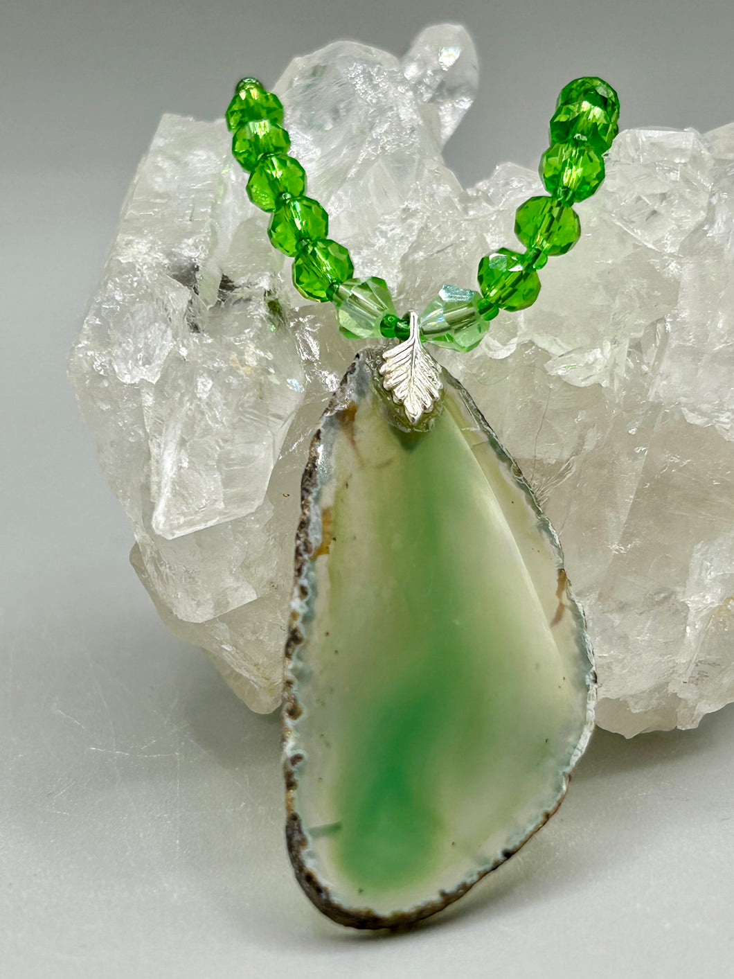 This Light Green Agate Geode Sliced Stone Chip Necklace is a statement piece for your jewelry collection. It features a bold green agate geode pendant surrounded by a glass bead necklace in varying shades of green. The silver leaf bail completes this chic, Boho-inspired necklace.  Measurements:   Necklace is 18” in length - pendant is 2 1/4” Height