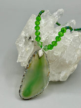Load image into Gallery viewer, This Light Green Agate Geode Sliced Stone Chip Necklace is a statement piece for your jewelry collection. It features a bold green agate geode pendant surrounded by a glass bead necklace in varying shades of green. The silver leaf bail completes this chic, Boho-inspired necklace.  Measurements:   Necklace is 18” in length - pendant is 2 1/4” Height
