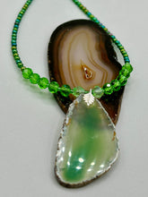 Load image into Gallery viewer, This Light Green Agate Geode Sliced Stone Chip Necklace is a statement piece for your jewelry collection. It features a bold green agate geode pendant surrounded by a glass bead necklace in varying shades of green. The silver leaf bail completes this chic, Boho-inspired necklace.  Measurements:   Necklace is 18” in length - pendant is 2 1/4” Height
