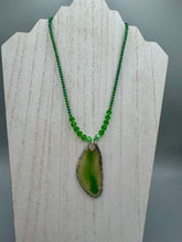 Load image into Gallery viewer, This Light Green Agate Geode Sliced Stone Chip Necklace is a statement piece for your jewelry collection. It features a bold green agate geode pendant surrounded by a glass bead necklace in varying shades of green. The silver leaf bail completes this chic, Boho-inspired necklace.  Measurements:   Necklace is 18” in length - pendant is 2 1/4” Height
