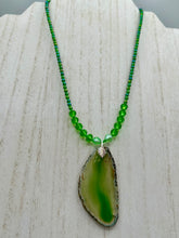Load image into Gallery viewer, This Light Green Agate Geode Sliced Stone Chip Necklace is a statement piece for your jewelry collection. It features a bold green agate geode pendant surrounded by a glass bead necklace in varying shades of green. The silver leaf bail completes this chic, Boho-inspired necklace.  Measurements:   Necklace is 18” in length - pendant is 2 1/4” Height
