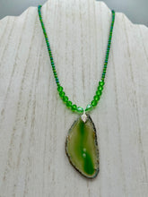 Load image into Gallery viewer, This Light Green Agate Geode Sliced Stone Chip Necklace is a statement piece for your jewelry collection. It features a bold green agate geode pendant surrounded by a glass bead necklace in varying shades of green. The silver leaf bail completes this chic, Boho-inspired necklace.  Measurements:   Necklace is 18” in length - pendant is 2 1/4” Height
