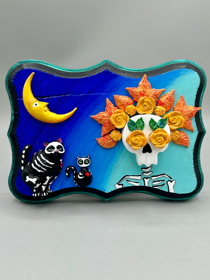 La Catrina y Gatita wall art is the purrfect hanging Day of the Dead art piece to add to your ofrenda. A tribute to an unbreakable bond and eternal love between cats and their guardians. Featuring a vibrant and intricate detail catrina and her floral crown accompanied by her beloved kitty cat. This Day of the Dead-inspired artwork adds a little touch of Mexican art to your home or office. A unique handcrafted gift for any cat lover. Measurements: H 5" x W 7” x D 1 1/2"