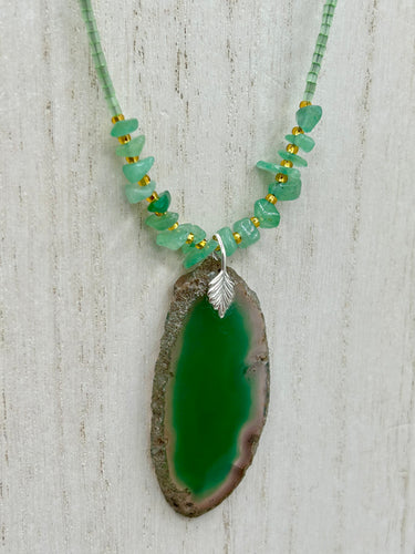 This Dark Green Agate Geode Sliced Stone Chip Necklace is a statement piece for your jewelry collection. It features a bold dark green agate geode pendant surrounded by a green aventurine, gold tone and green glass beaded necklace. The silver leaf bail completes this chic, Boho-inspired necklace.  Measurements:   Necklace is 19” in length - pendant is 2 1/4” Height