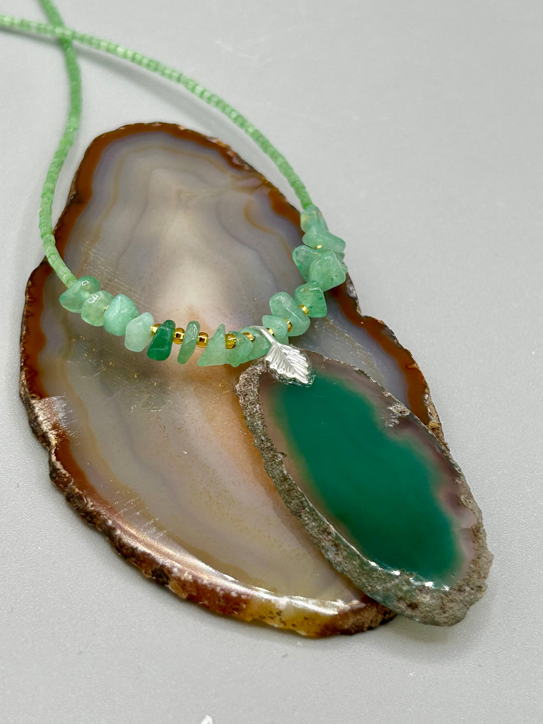 This Dark Green Agate Geode Sliced Stone Chip Necklace is a statement piece for your jewelry collection. It features a bold dark green agate geode pendant surrounded by a green aventurine, gold tone and green glass beaded necklace. The silver leaf bail completes this chic, Boho-inspired necklace.  Measurements:   Necklace is 19” in length - pendant is 2 1/4” Height