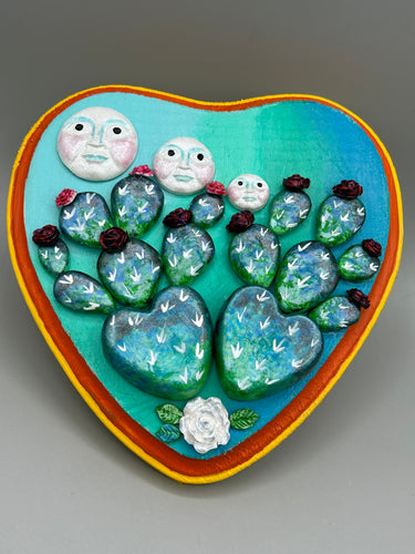 This handcrafted polymer clay wall art features a heart-shaped wood frame complimented with three smiling moon motifs.  Each moon symbolizing each of the realms of heavens, earth, and underworld. The complimentary color palette consists of cool sky hues, from a bright turquoise to a dark royal blue. The piece is further enhanced with hand sculpted cacti plants with blooming light and dark pink flowers.  The vibrant red and orange frame complements the cool-toned artwork.   H 7