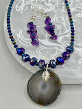 Load image into Gallery viewer, These Lapis Stone Chip Beaded Hoop Earrings feature a dark blue-tone pressed glass leaf charm with an iridescent shimmer finish showcasing hues of gold, purple, blue and pink. The earrings display natural lapis stone chips and royal blue with gold glass beads.  Measurements: 1 1/2&quot; length
