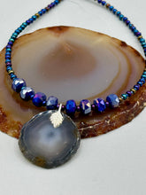 Load image into Gallery viewer, This Dark Gray-blue Agate Geode Sliced Stone Chip Necklace is a unique, eye-catching accessory to elevate your jewelry collection. The bold pendant features a striking gray-blue agate geode surrounded by eye-catching, shimmery dark-purple glass beads with beautiful iridescence that reflects hues of blue, pink, gold, and green. It&#39;s a perfect boho chic accent to update any look.   Measurements:   Necklace is 18” in length - pendant is 1 1/4” Height
