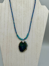 Load image into Gallery viewer, This Dark Blue Agate Geode Sliced Stone Chip Necklace is a luxurious addition to any jewelry collection. It features bold, eye-catching dark blue agate geode in an earthy outline, suspended on a unique glass beaded necklace that contains a mixture of blue and purple glass beads with a beautiful iridescence. This necklace is the perfect boho chic accent to take any look to the next level.   Measurements:    Necklace is 18” in length - pendant is 1 3/4” Height
