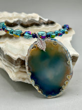 Load image into Gallery viewer, This Dark Blue Agate Geode Sliced Stone Chip Necklace is a luxurious addition to any jewelry collection. It features bold, eye-catching dark blue agate geode in an earthy outline, suspended on a unique glass beaded necklace that contains a mixture of blue and purple glass beads with a beautiful iridescence. This necklace is the perfect boho chic accent to take any look to the next level.   Measurements:    Necklace is 18” in length - pendant is 1 3/4” Height
