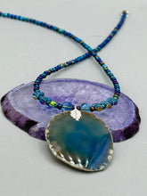 Load image into Gallery viewer, This Dark Blue Agate Geode Sliced Stone Chip Necklace is a luxurious addition to any jewelry collection. It features bold, eye-catching dark blue agate geode in an earthy outline, suspended on a unique glass beaded necklace that contains a mixture of blue and purple glass beads with a beautiful iridescence. This necklace is the perfect boho chic accent to take any look to the next level.   Measurements:    Necklace is 18” in length - pendant is 1 3/4” Height

