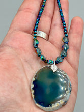 Load image into Gallery viewer, This Dark Blue Agate Geode Sliced Stone Chip Necklace is a luxurious addition to any jewelry collection. It features bold, eye-catching dark blue agate geode in an earthy outline, suspended on a unique glass beaded necklace that contains a mixture of blue and purple glass beads with a beautiful iridescence. This necklace is the perfect boho chic accent to take any look to the next level.   Measurements:    Necklace is 18” in length - pendant is 1 3/4” Height
