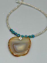 Load image into Gallery viewer, This handcrafted necklace is perfect for anyone who loves an earthy, bohemian look. Crafted with a unique strand of gold and clear beading, blue glass beads with iridescent hues, and natural banded agate geode, this piece adds style and luxury to any ensemble. Measurements:     Necklace is 18” in length - pendant is 2” Height 
