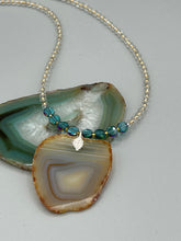 Load image into Gallery viewer, This handcrafted necklace is perfect for anyone who loves an earthy, bohemian look. Crafted with a unique strand of gold and clear beading, blue glass beads with iridescent hues, and natural banded agate geode, this piece adds style and luxury to any ensemble. Measurements:     Necklace is 18” in length - pendant is 2” Height 
