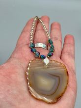 Load image into Gallery viewer, This handcrafted necklace is perfect for anyone who loves an earthy, bohemian look. Crafted with a unique strand of gold and clear beading, blue glass beads with iridescent hues, and natural banded agate geode, this piece adds style and luxury to any ensemble. Measurements:     Necklace is 18” in length - pendant is 2” Height 
