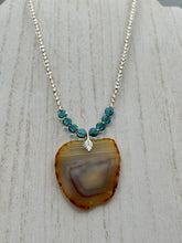 Load image into Gallery viewer, This handcrafted necklace is perfect for anyone who loves an earthy, bohemian look. Crafted with a unique strand of gold and clear beading, blue glass beads with iridescent hues, and natural banded agate geode, this piece adds style and luxury to any ensemble. Measurements:     Necklace is 18” in length - pendant is 2” Height 
