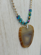 Load image into Gallery viewer, This handcrafted necklace is perfect for anyone who loves an earthy, bohemian look. Crafted with a unique strand of gold and clear beading, blue glass beads with iridescent hues, and natural banded agate geode, this piece adds style and luxury to any ensemble. Measurements:     Necklace is 18” in length - pendant is 2” Height 

