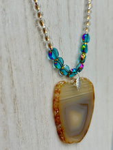 Load image into Gallery viewer, This handcrafted necklace is perfect for anyone who loves an earthy, bohemian look. Crafted with a unique strand of gold and clear beading, blue glass beads with iridescent hues, and natural banded agate geode, this piece adds style and luxury to any ensemble. Measurements:     Necklace is 18” in length - pendant is 2” Height 

